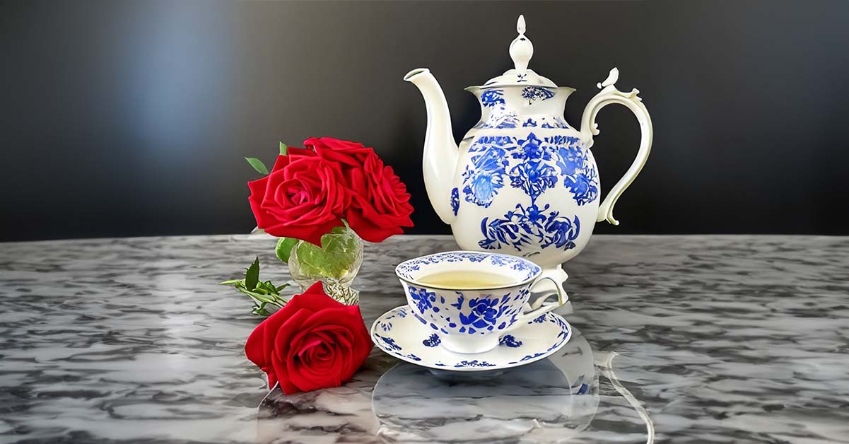 A crystal-clear teacup filled with steaming ginger tea sits atop a polished black marble counter. The pristine white china cup has delicate blue motifs around the rim. A matching china teapot and sugar bowl complete the set, alongside a vase holding two red roses. The elegant yet understated china symbolizes refinement through everyday connection. A tea set built to endure, complementing a marriage that has matured into resilient commitment. The steaming cup represents warmth and comfort between partners. The roses signify romance still blooming after two years of wedded bliss.