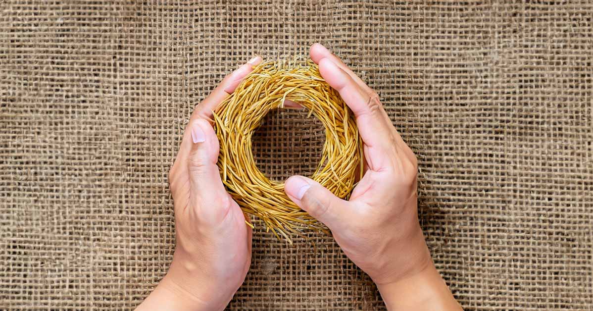 A pair of tan, weathered hands tenderly holding a neatly woven ring made of golden wheat straw against a rustic burlap backdrop. The interlaced straw symbolizes the joining of two lives into one unbreakable union during the humble, flexible early years of marriage. A few pieces of straw are peeking out, reminiscent of rough edges and imperfections. But bound tightly together, the straw is transformed into something beautiful, sturdy and purposeful. The loving hands provide a protective embrace around the straw ring, representing comfort through life's storms. Sunlight streams in, suggestive of the brighter phases still to come.